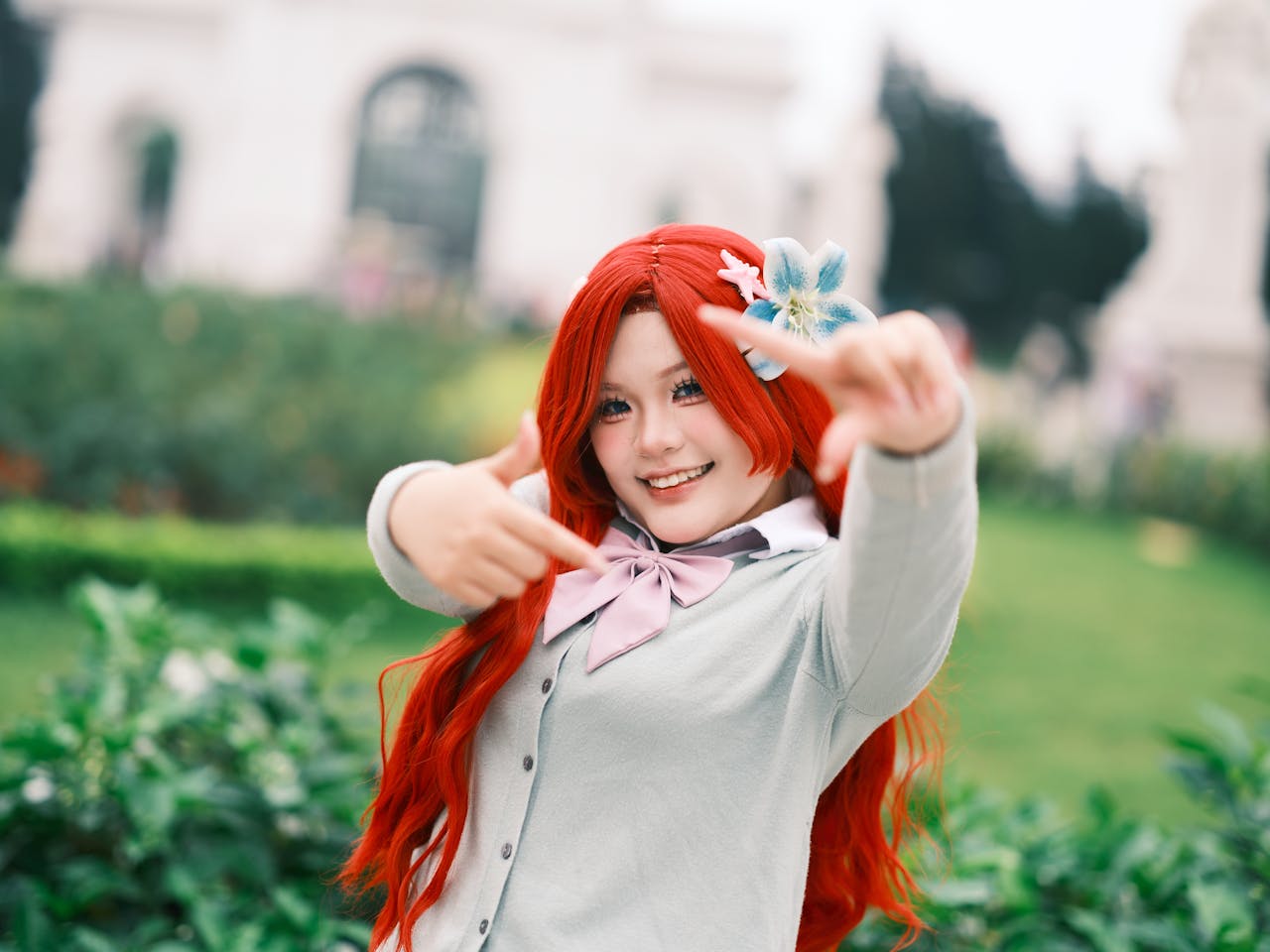 Cheerful cosplayer with red hair and floral accessory posing outdoors in a garden, Hanoi, Vietnam.